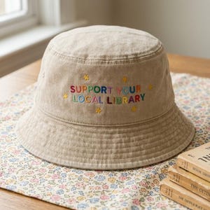 May include: Beige bucket hat with the embroidered text "SUPPORT YOUR LOCAL LIBRARY" in colorful thread. The hat has a structured brim and is made of a textured fabric. Small star designs are embroidered around the text.