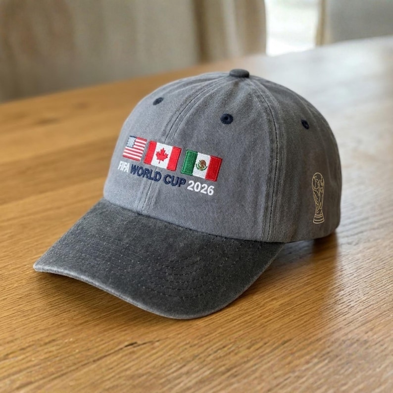 May include: Gray baseball cap with a curved brim. The cap features embroidered flags of the USA, Canada, and Mexico, along with the text "FIFA WORLD CUP 2026". A gold-colored FIFA World Cup trophy is on the side.
