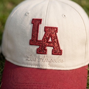 May include: A beige baseball cap featuring the letters LA in red glitter on the front crown. Below the letters, the words Los Angeles are embroidered in a matching beige script. The cap has a solid red brim and stitched eyelets on the crown.