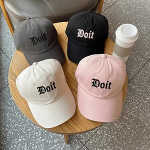 May include: Four baseball caps in gray, black, white, and pink, each with the word "Doit" embroidered in a gothic font. The caps are arranged on a wooden table, with a disposable coffee cup visible.