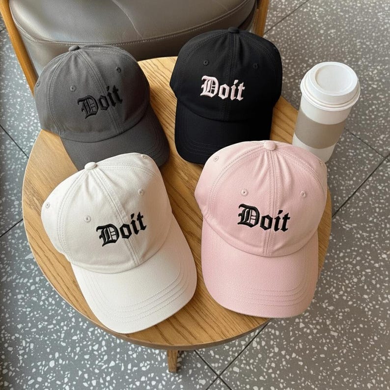 May include: Four baseball caps in gray, black, white, and pink, each with the word "Doit" embroidered in a gothic font. The caps are arranged on a wooden table, with a disposable coffee cup visible.