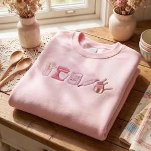 May include: A light pink crewneck sweatshirt featuring embroidered kitchen-themed icons in shades of pink and white. The design includes a utensil holder, a stand mixer, a bag labeled flour, a rolling pin, and a set of measuring cups.
