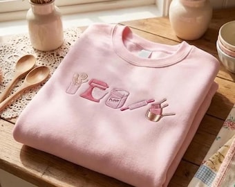 Peut inclure: Un sweat-shirt à col rond rose clair orné d'icônes de cuisine brodées dans des tons de rose et de blanc. Le motif comprend un porte-ustensiles, un batteur sur socle, un sac étiqueté flour, un rouleau à pâtisserie et des tasses à mesurer.