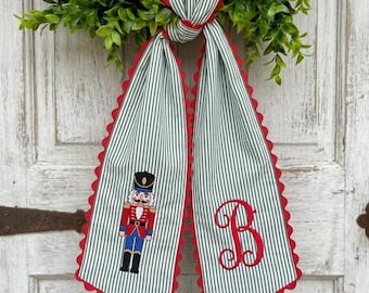 May include: A decorative fabric hanging featuring a green and white pinstripe pattern with red scalloped edges. One side displays an embroidered nutcracker figure in a red uniform, while the other side features a large, red embroidered letter B.