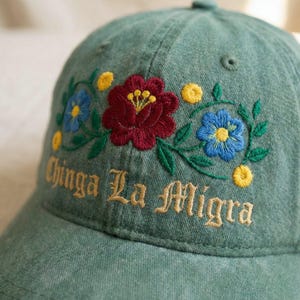 May include: A green baseball cap with embroidered floral designs in red, blue, and yellow. The text "Chinga La Migra" is embroidered in gold thread on the front of the cap. The cap has a curved brim and a button on top.