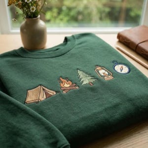 Peut inclure: Un sweat-shirt &agrave; col rond vert fonc&eacute; orn&eacute; d'une rang&eacute;e d'ic&ocirc;nes brod&eacute;es sur le th&egrave;me du camping, comprenant une tente beige, un feu de camp, un sapin vert, une lanterne marron et une boussole bleue et blanche sur la poitrine.