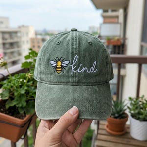May include: Green baseball cap with a curved brim. The cap features an embroidered bee and the word "kind" in white script. The cap is made of a soft, worn material.