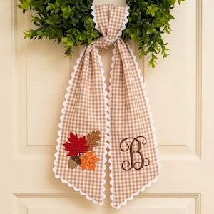 May include: A tan and white gingham fabric ribbon with scalloped white edges, featuring an embroidered design of red, orange, and brown autumn leaves with an acorn on one side and a large, dark brown embroidered letter B on the other side.