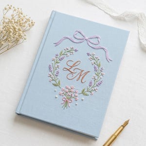 May include: A light blue hardcover journal featuring an embroidered design on the front cover. The embroidery includes a lavender bow, a floral wreath with small pink and purple flowers, and the copper-colored cursive initials LM in the center.