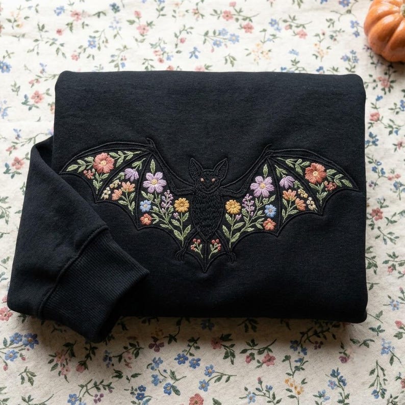 Beautifully stitched across the chest of an ultra-soft crewneck pullover, this detailed design features a nocturnal bat whose outstretched wings are filled with vibrant embroidered wildflowers—including pastel daisies, lavender sprigs, and lush green foliage. Available in deep forest green and classic black, this cozy sweater seamlessly blends dark gothic charm with whimsical botanical beauty for the ultimate spooky season staple!