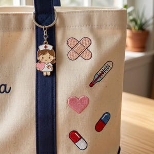 May include: Cream-colored tote bag with navy blue handles and accents. A nurse keychain hangs from the handle. Embroidered patches of a bandage, thermometer, heart, and pills decorate the bag. The bag is on a wooden surface.