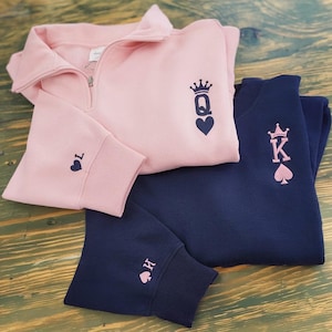 May include: Two sweatshirts, one pink and one navy blue, are displayed. The pink sweatshirt has a quarter-zip neckline and a "Queen" graphic with a heart. The navy blue sweatshirt has a "King" graphic with a spade. Both have card suit symbols.