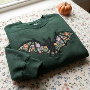 Peut inclure: Un sweat-shirt &agrave; col rond vert for&ecirc;t fonc&eacute; orn&eacute; d'une grande broderie repr&eacute;sentant une chauve-souris noire aux ailes d&eacute;ploy&eacute;es. Les ailes de la chauve-souris sont remplies de broderies florales complexes et color&eacute;es dans des tons rose, jaune, bleu et blanc.