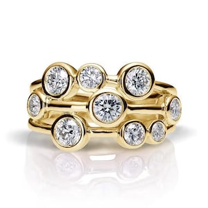 Puede incluir: Una pila de tres anillos de oro, cada uno con múltiples gemas redondas y transparentes. Los anillos tienen una superficie pulida y reflectante, y las gemas están engastadas en biseles. El diseño es moderno y elegante.