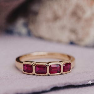 May include: A gold ring featuring four rectangular, emerald-cut red rubies set in a horizontal row. The stones are held in a bezel setting, creating a smooth, continuous band design. The metal has a polished finish, highlighting the vibrant red color.