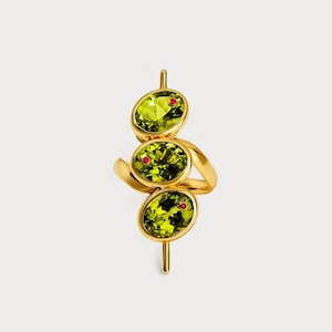 May include: A gold ring featuring three oval-shaped, light green gemstones with small red accents. The ring's design is a unique, modern take on a cocktail ring, with a vertical arrangement of the stones.