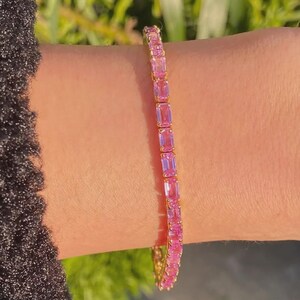 May include: A gold bracelet featuring a row of rectangular pink gemstones. The bracelet is worn on a wrist, with a black sleeve visible in the background. The gemstones are a vibrant pink color.