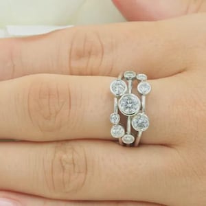 Pode incluir: Um anel de prata com múltiplas pedras preciosas redondas e transparentes. O anel apresenta um design único com pedras de diferentes tamanhos, criando um efeito de aglomerado. O anel é usado em uma mão, mostrando sua aparência brilhante.