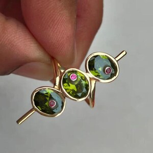 Op de afbeelding: Een gouden ring met drie ovale groene edelstenen, elk met een klein roze accent. De ring heeft gouden staven die zich van elke edelsteen uitstrekken. De ring wordt door een hand omhoog gehouden.