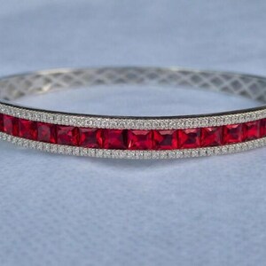 Puede incluir: Brazalete plateado con una fila de piedras preciosas rojas cuadradas y brillantes. El brazalete está bordeado por una fila de pequeñas piedras preciosas transparentes. El cierre es visible en el lado izquierdo.