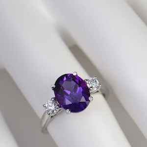 May include: A silver ring with a large, oval-cut, deep purple amethyst gemstone as the centrepiece, with two smaller, round, clear gemstones either side. The ring is set against a white background.