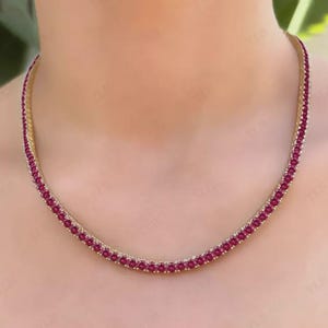 May include: A gold-toned necklace featuring a continuous row of small, round, deep red gemstones. The necklace is designed to sit close to the neckline, showcasing the vibrant color of the stones. The necklace is a piece of jewelry.