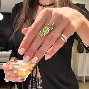 May include: A hand displays three gold rings with oval green gemstones and a silver ring with three clear stones. The hand also holds a martini glass with olives. The nails are painted a light pink.
