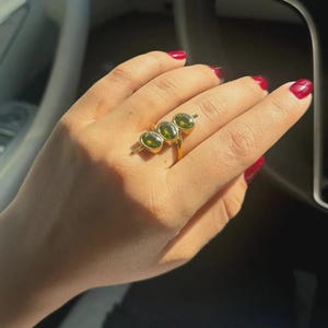 May include: A gold ring featuring three oval-cut, olive-green gemstones. The ring's band is a simple, polished gold, and the gemstones are set horizontally across the finger. The ring is shown on a hand with red nail polish.