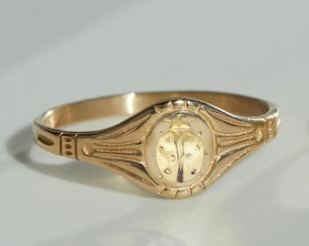 Può includere: Un anello dorato con un design dettagliato di coccinella al centro. L'anello ha una forma ovale con dettagli intricati attorno alla coccinella e lungo la fascia. L'anello è probabilmente in oro o metallo dorato.
