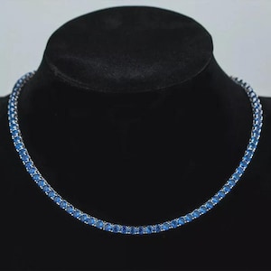 May include: A tennis necklace featuring a continuous line of round, brilliant-cut blue gemstones. The necklace is displayed on a black jewelry bust, showcasing the vibrant blue stones against a silver-toned metal setting. The gemstones are evenly spaced.