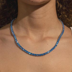 May include: A sparkling blue gemstone necklace. The necklace is composed of numerous round, light blue gemstones set closely together, creating a continuous line of color. The necklace is worn around the neck, reflecting light and adding a touch of elegance.
