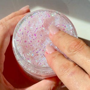 Pode incluir: Um recipiente redondo e transparente cheio de contas rosa, brilhantes e iridescentes. O recipiente é segurado pelas mãos de uma pessoa. O produto parece ser um item cosmético, como um esfoliante corporal ou um produto de banho.
