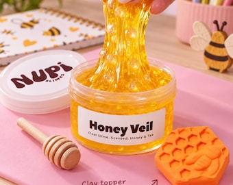 May include: A clear yellow slime with embedded beads in a round plastic container labeled Honey Veil Clear Slime Scented Honey & Tea. The container has a white lid with the text Nupi Slimes. A wooden honey dipper and an orange bee-shaped clay topper are nearby.