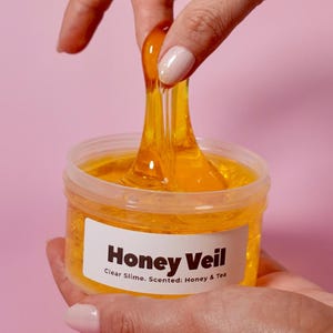 May include: A jar of clear, golden-yellow slime labeled "Honey Veil." The slime is being stretched, revealing its viscous texture. The label indicates it's scented with honey and tea. The background is a solid pink.