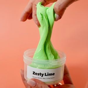以下が含まれることがあります： 鮮やかな緑色のスライムが入った透明なプラスチック容器。「Zesty Lime」とラベルされています。スライムが伸ばされ、その質感が現れています。ラベルには「Butter Slime. Key Lime Scent」とも記載されています。