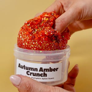 May include: A clear plastic container labeled "Autumn Amber Crunch" holds a vibrant, textured slime. The slime is a deep orange color, speckled with glitter and small, iridescent beads. The label also mentions a pumpkin spice scent.
