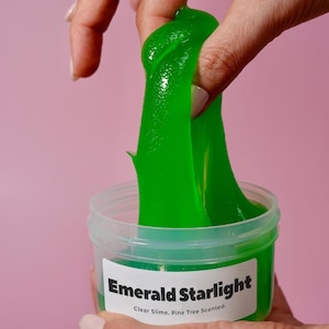 May include: A clear plastic container filled with bright green slime. The slime is being pulled up and out of the container, revealing its glossy texture. The label reads "Emerald Starlight, Clear Slime, Pine Tree Scented."