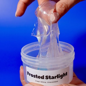 May include: A clear, unscented slime labeled "Frosted Starlight" is held up, revealing its translucent, shimmering texture. The slime is in a clear plastic container against a vibrant blue background. The text "Clear Slime. Unscented." is printed on the label.