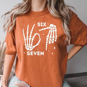 An orange t-shirt with a graphic design featuring skeletal hands and the text "SIX SEVEN" printed on it.