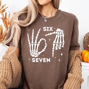 A woman wearing a brown t-shirt with a graphic design of skeletal hands and the text "SIX SEVEN" printed on it.