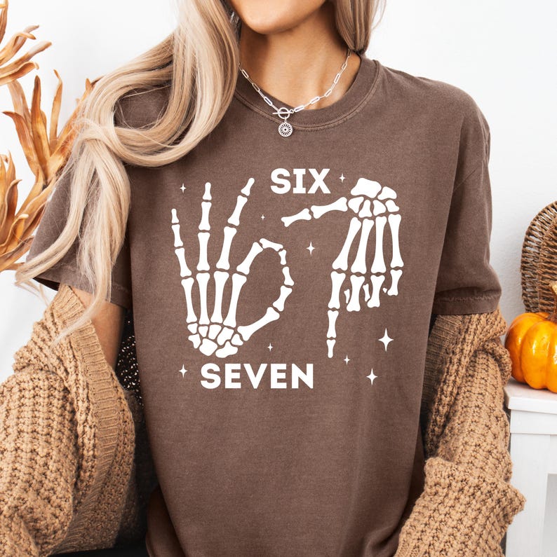 A woman wearing a brown t-shirt with a graphic design of skeletal hands and the text "SIX SEVEN" printed on it.