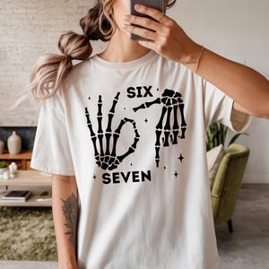 A person wearing a white t-shirt with a graphic design featuring skeletal hands and the text "SIX SEVEN" printed on it.
