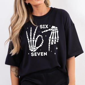 A woman wearing a black t-shirt with a graphic design featuring skeletal hands and the text "SIX SEVEN" printed on it.