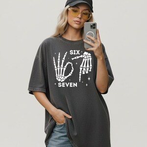 A woman wearing a black cap and glasses is taking a selfie with her phone, holding it up to her face. She is wearing a gray t-shirt with a graphic design and text printed on it.