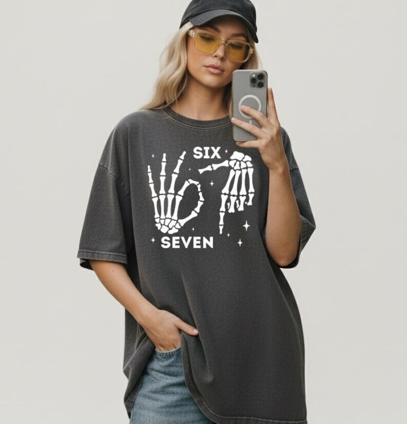A woman wearing a black cap and glasses is taking a selfie with her phone, holding it up to her face. She is wearing a gray t-shirt with a graphic design and text printed on it.
