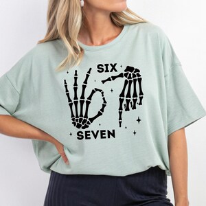 A woman wearing a light green t-shirt with a graphic design featuring a skeleton hand and the text "SIX SEVEN" printed on it.