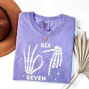 a purple t-shirt with a skeleton design and the text "Six Seven" printed on it, along with a brown hat and a fan.