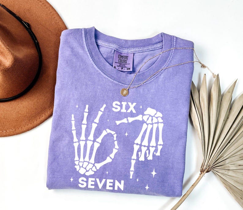 a purple t-shirt with a skeleton design and the text "Six Seven" printed on it, along with a brown hat and a fan.