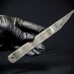 May include: A Damascus steel knife with a textured blade and pointed tip. The knife has a serrated edge near the handle and the word "TAYZER" is etched on the blade. The knife is held against a dark background.