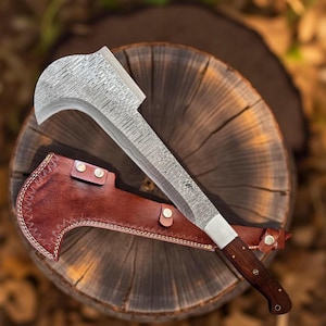 May include: A large, curved blade machete with a patterned steel blade and a dark wooden handle. The machete rests on a wooden surface, with a brown leather sheath. The blade has a white section near the handle.